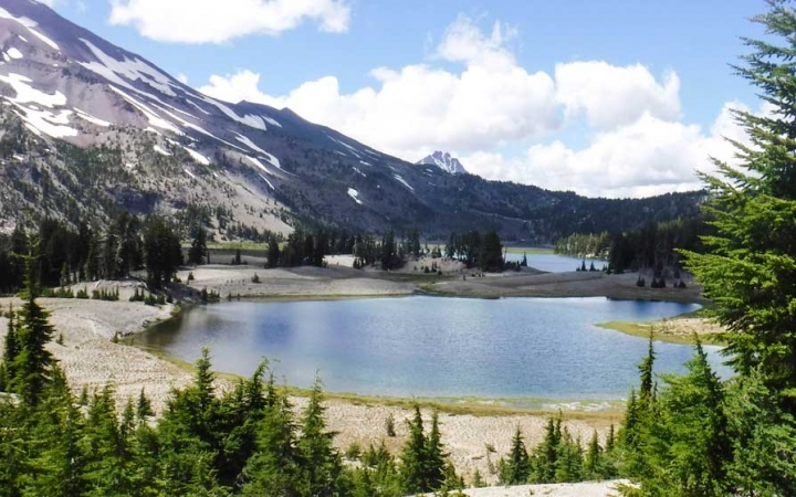 An alpine lake rests among evergreen trees and gray mountains dotted with snow
